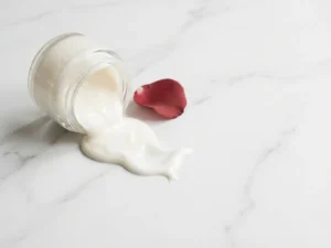 Glazen potje huidcrème gekanteld op wit marmer met gemorste crème en rood rozenblaadје, zachte natuurlijke belichting