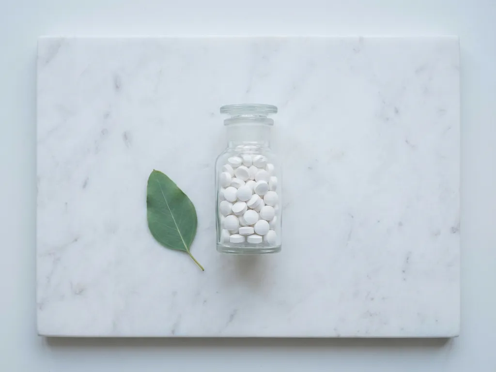 Glazen medicijnflesje met witte tabletten op wit marmeren oppervlak met groen blad accent