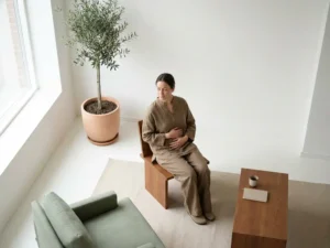 Persoon zit vredig in lichte medische consultatieruimte met handen op torso, plant bij raam, minimalistisch interieur