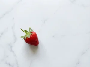 Enkele rode aardbei op wit marmeren oppervlak met zachte natuurlijke belichting en minimalistische compositie