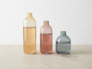 Drie glazen vazen op stenen ondergrond gevuld met amber, roze en grijsblauw vloeistof, één gebarsten en lekkend.