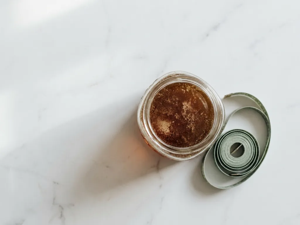 Glazen pot donkergele kristalliserende honing op wit marmer met een opgerolde meetlint, bovenaanzicht.